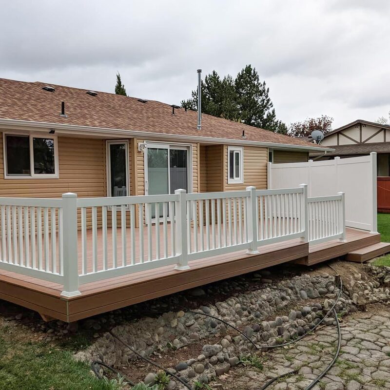 This professional backyard deck installation combines modern materials for long-lasting beauty and function. A multi-level composite deck in a natural wood-grain brown finish creates a large outdoor living area. The deck is enclosed by a stark white vinyl railing system with vertical balusters.