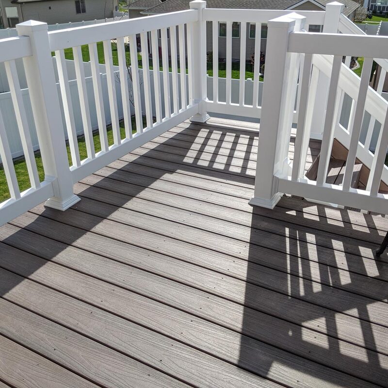 Enhance your outdoor living with a composite deck built for durability and comfort. This installation, viewed from above, features low-maintenance gray-brown composite planks. A robust white vinyl railing system with vertical balusters and decorative post caps defines the space. Integrated composite stairs lead down to a lower patio area. The project is completed in a suburban setting, adjacent to a green lawn and a white vinyl privacy fence, showcasing a finished patio chair with a cushion for a move-in-ready feel. The structure overlooks a neighborhood with other houses.