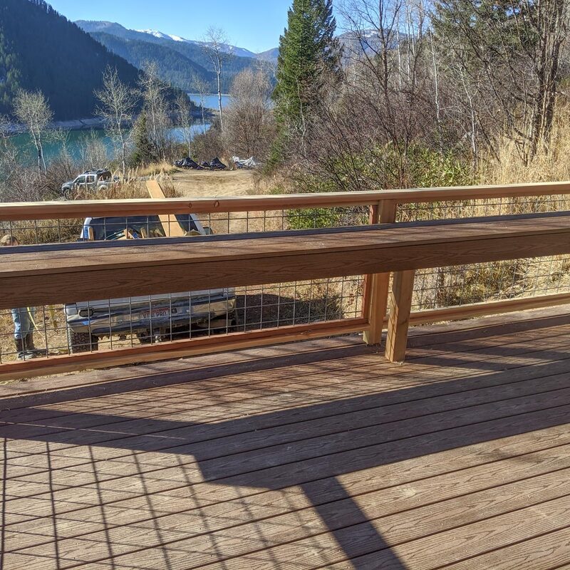 Embrace rustic charm with this custom composite deck by DeckPro LLC. Designed to blend perfectly with the natural surroundings and the building's corrugated siding, it features durable brown composite decking. The unique wood and industrial wire mesh railing offers safety without blocking the view, alongside a convenient built-in bench for relaxing outdoors.