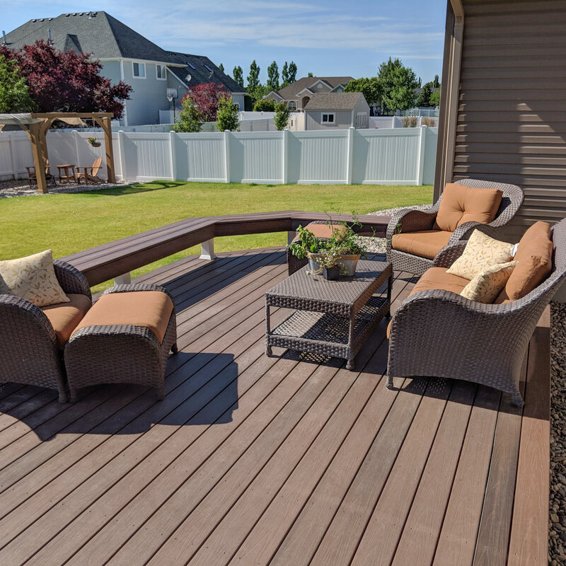 Create the ultimate space for relaxing and entertaining with this custom composite deck by DeckPro LLC. This expansive deck features low-maintenance, dark brown composite decking boards and a unique built-in perimeter bench that doubles as safe railing. Furnished with comfortable wicker lounge seating and a coffee table, it’s the perfect spot to enjoy your meticulously manicured lawn and peaceful suburban backyard.