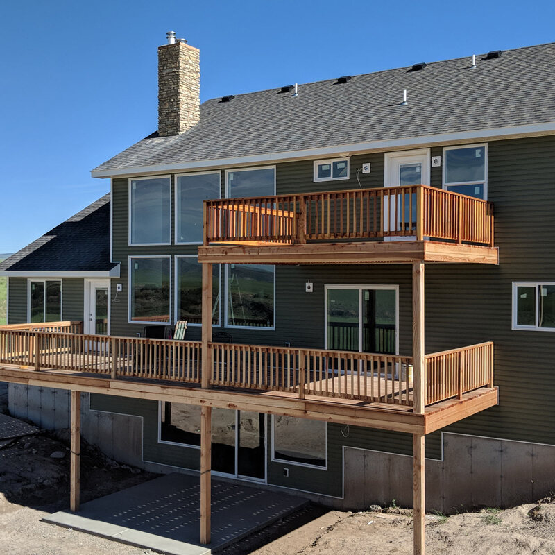 Enhance your new construction with a stunning multi-level natural wood deck by DeckPro LLC. This custom design features an upper-level balcony accessible from the master suite and a expansive lower-level deck perfect for entertaining, all with safe and stylish wood railings. The warm wood tones create a perfect contrast with the deep olive green siding, integrating beautifully with the stone veneer chimney and surrounding open landscape.