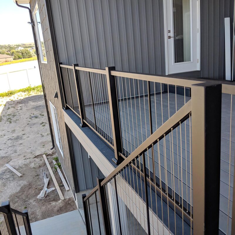 A sophisticated elevated deck is showcased, featuring a contemporary design. The key highlight is the black aluminum post and cable railing system, which provides a sleek, modern look while maximizing the expansive field and residential views. The grey composite decking ensures longevity and low maintenance. This design integrates seamlessly with the vertical grey siding of the house.