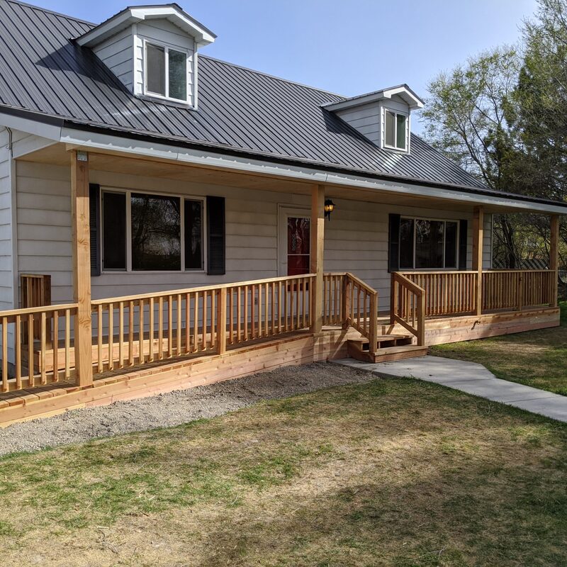 Extend your multi-level outdoor living with this dramatic multi-zone composite raised deck transformation by DeckPro LLC. Pictured on a beautiful partly cloudy day, this sophisticated structure features expansive gray and brown composite planks paired with sleek black vertical perimeter railings. This sturdy retreat transitions via extensive massive wide staircases with contrasting risers to a stamped concrete lower level and a beautifully landscaped rock garden featuring natural boulder rocks on a gravel bed. Direct access point via multiple existing glass doors and a large massive set of connecting stairs allow for effortless entertaining, while integrated recessed ceiling lighting in the covered upper area ensures evening use. Defining functional zones perfect for both relaxation and large-scale entertaining, this unique tranquilidad sanctuary maximizes spectacular multi-directional natural views.