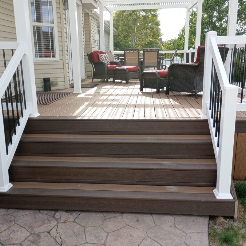 Create the perfect backyard retreat with this custom low-maintenance composite deck by DeckPro LLC. Featuring rich brown decking, a stylish white attached pergola for dappled shade, and modern white railings with black metal balusters, this outdoor space is ready for relaxing or entertaining. The wide stairs lead down to a beautiful stamped concrete patio.