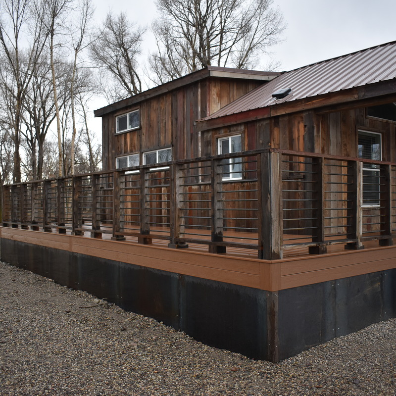A wide exterior shot showcasing a complete rustic deck installation by DeckPro LLC on a custom wood-sided cabin. The project features highly durable brown composite decking with matching fascia boards, transitioning into a solid, dark lower skirt to protect the understructure. The custom railing, built with dark timber posts and horizontal metal bars, delivers a striking mountain-cabin aesthetic that blends seamlessly with the rural environment.