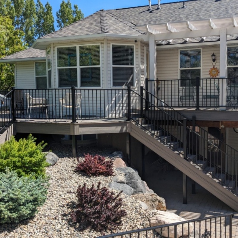 Extend your multi-level outdoor living with this sophisticated composite deck project by DeckPro LLC. This functional retreat features massive connecting stairs leading down to a beautifully integrated landscape with various bushes and large natural boulder rocks on a gravel bed. Perfect for relaxation or large-scale entertaining, this sturdy retreat transitions smoothly from defined sun and shade zones, utilizing expansive space under a covered pergola and optimizing multi-directional natural views. We installed high-quality composite decking planks in a clean pattern and paired them with safe and sophisticated dark vertical perimeter railings for defined zones that maximize multi-directional views. Pictured on a beautiful partly cloudy day, this backyard transformation creates a tranquil sanctuary perfect for large properties. Recessed lighting allows for extended evening use.