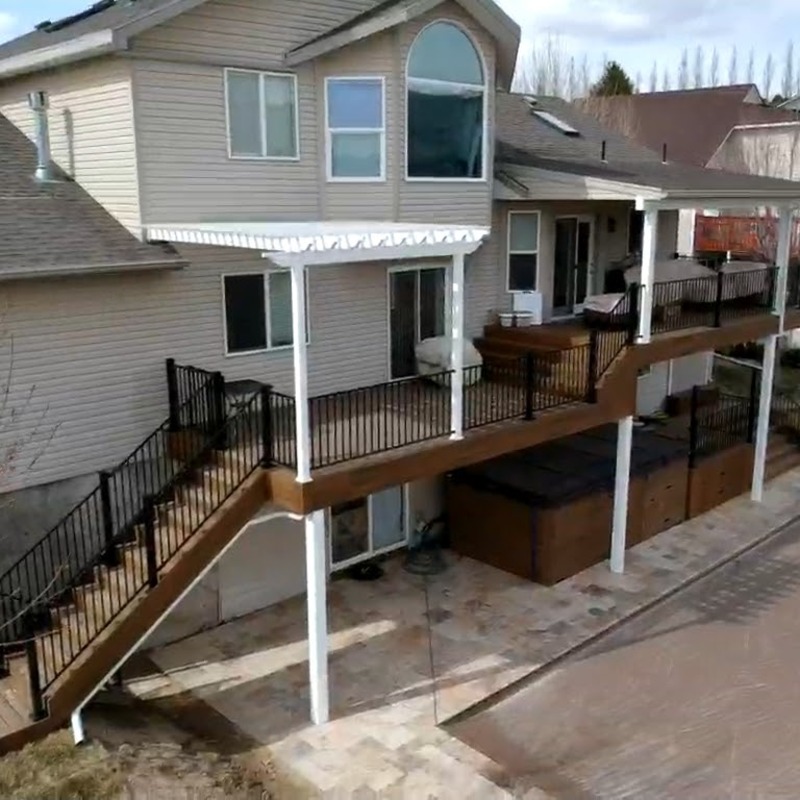 Extend your multi-level outdoor living with this expansive composite deck and patio transformation by DeckPro LLC. This functional retreat seamlessly connects a multi-story home with rough grading and rough landscaping, pictured during a transitional construction phase on a beautiful day with distinct shadows cast across the planks, and turns it into a sophisticated, multi-functional sanctuary perfect for both relaxation and large-scale entertaining under prominent covered upper area.