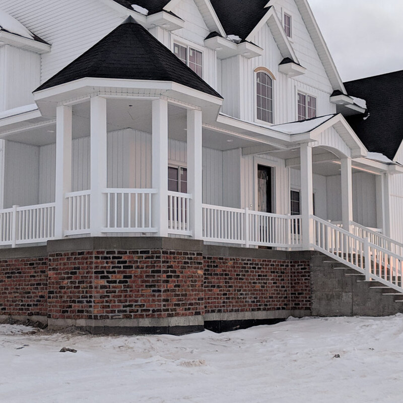 A detailed view of a newly constructed custom home in a snowy Utah landscape. This project features a large, wrap-around front porch with clean, maintenance-free white vinyl railings that define the space. The railing system includes vertical pickets, top and bottom rails, and sturdy posts. The porch is elevated and supported by a striking foundation base combining a prominent red brick skirt with visible concrete columns, adding architectural texture and stability. Concrete steps with matching white vinyl handrails lead up to the front entrance. The white siding of the house provides a seamless backdrop, under a multi-gabled black roof line.