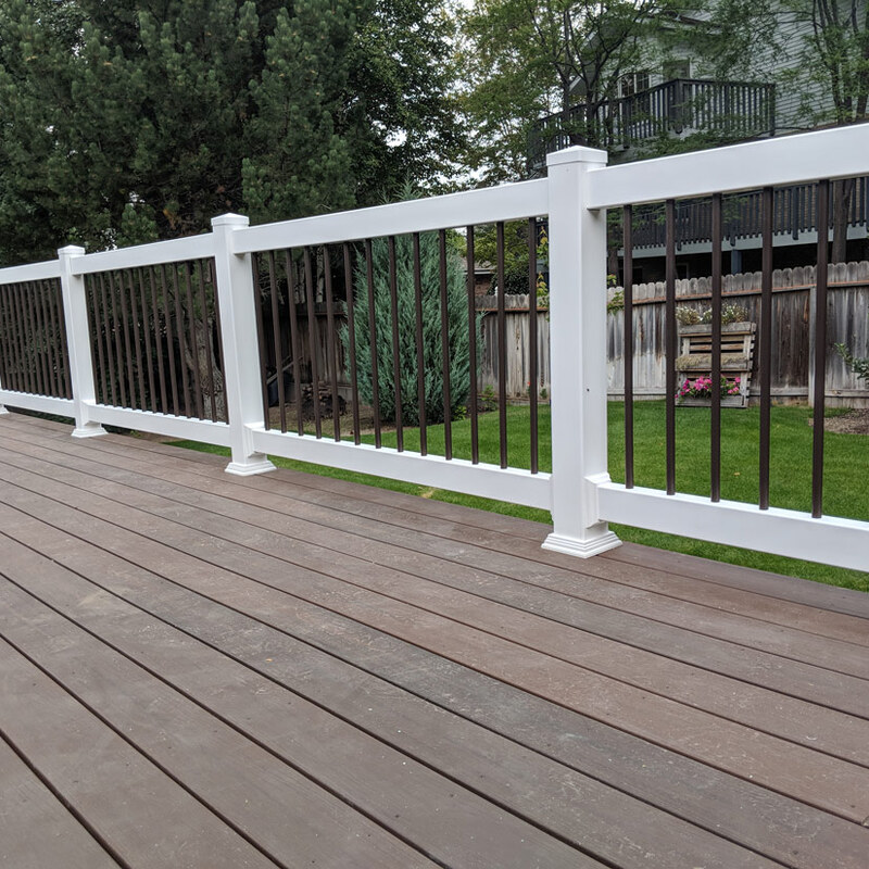 This elevated composite deck installation features durable, dark wood-grain composite planks that create a sleek, low-maintenance surface. The perimeter is enclosed by a premium hybrid railing system: clean white vinyl posts and top/bottom rails are contrasted beautifully by sleek, dark bronze vertical metal balusters. This design provides safety without sacrificing the view of the lush, green lawn and surrounding nature in this idaho backyard.